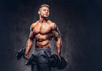 Fototapeta na wymiar A handsome shirtless tattooed bodybuilder with stylish haircut and beard, wearing sports shorts, posing in a studio. Isolated on a dark background