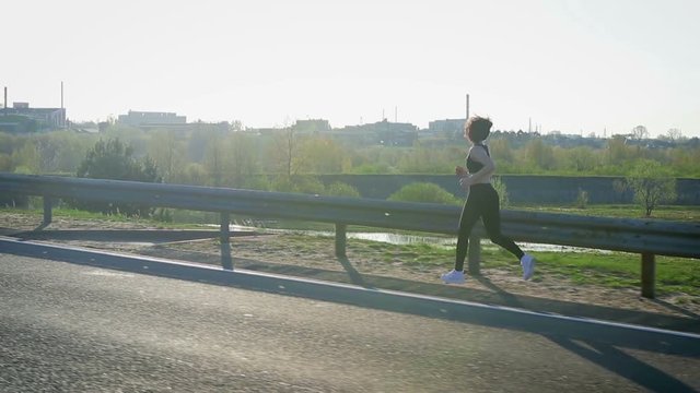 Young pretty sportswoman is engaged in fitness in nature. The girl runs along the road. Steadicam shot