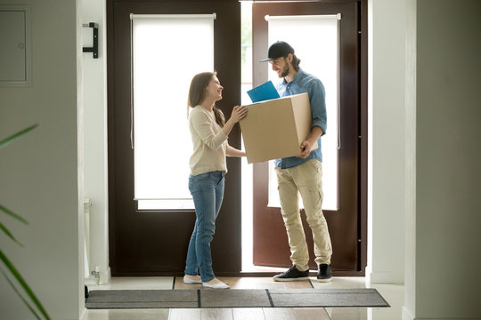 Happy young woman receiving parcel from smiling courier at home, delivery man holding carrying package giving cardboard box to customer receiver standing at doorway, door delivery service concept