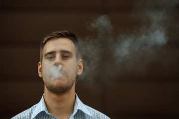 Obraz premium Vape man. Portrait of a handsome young white guy with modern haircut in in checkered sleeveless shirt vaping an electronic cigarette opposite dark background in the spring. Lifestyle.