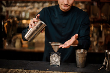 Fototapeta na wymiar Bartender pouring fresh drink from a shaker into a glass using strainer