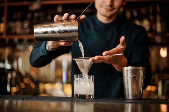 Smiling bartender pouring fresh drink from a shaker into a glass using strainer