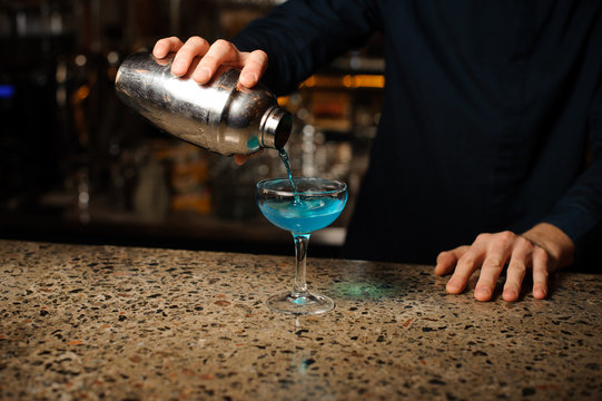 Barman hand pouring fresh drink with blue liquor from a shaker into a glass using strainer