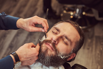 Bearded male sitting in an armchair in a barber shop while hairdresser shaves his beard with a dangerous razor.