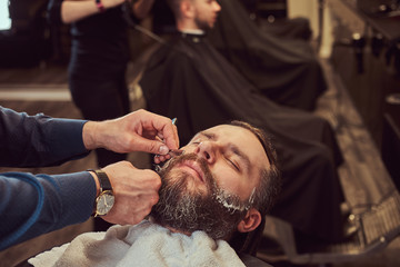 Bearded male sitting in an armchair in a barber shop while hairdresser shaves his beard with a dangerous razor.