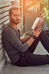 Fototapeta premium Handsome stylish redhead student in casual clothes, sitting on a floor at a home and holds a book, looking at a camera.