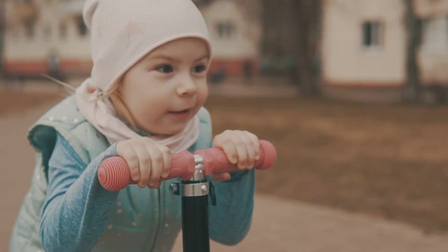 Little girl goes for ride on pink scooter at sidewalk in slow motion.