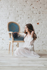 A young girl in a white dress with a bouquet of lavender sits on a wooden floor near the chest.