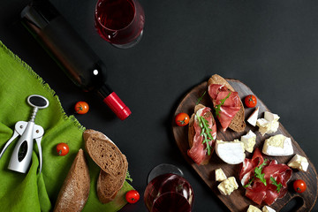 Red wine, cheese, cherry tomato, bread and prosciutto on wooden board over black backdrop, top...