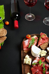 Prosciutto, salami, baguette slices, tomatoes and nutson rustic wooden board, two glasses of red wine over black background