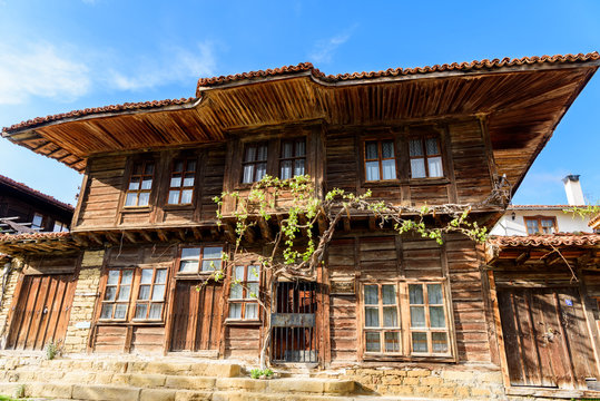 Low angle view of the front side of old traditional rustic house made of wood and covered by grape vine with eaves on top against blue sky, Zheravna, Bulgaria