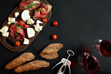 Prosciutto, salami, baguette slices, tomatoes and nutson rustic wooden board, two glasses of red wine over black background