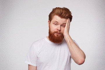 Indoor shot of bearded ginger male feels tired, keeps his hand on cheek, looks  forworn and aweary....