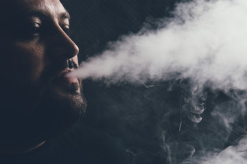 Man vape e-cigarette with e-liquid, close-up, breathes out large cloud of steam or vapor on dark...