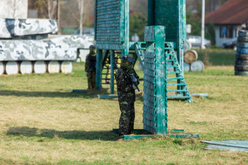 Boy in the camouflage holds a paintball gun  in one hand and protective helmet , standing on the field with group of players on the background