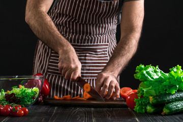 Young home cook man in apron slicing carrot with kitchen knife.