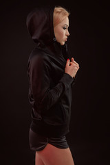 Fototapeta na wymiar Beautiful seductive sporty girl with the slim sexy figure in the black hoodie, sport underwear and little black shorts is posing in the studio