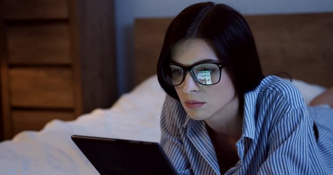 Close up of the young Cacasian woman with long dark hair lying on the bed on the stomach, watching something on the tablet device late at night. Indoors