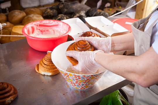 A close-up of a female baker in protective gloves  prepares buns cinnapons and pours cream on them in bakery