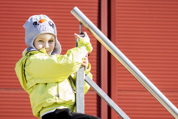 Portrait of Caucasian boy in hat and jacket on railing of staircase. boy holds onto railing and smiles. Children's joy and happiness on the boy's face.