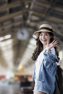 Asian backpack traveler woman smiling and waving hand at train station platform, summer holiday travelling or young tourist concept
