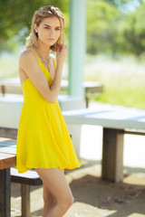 Attractive young blonde Caucasian woman in thin yellow sun dress posing outside on wooden table