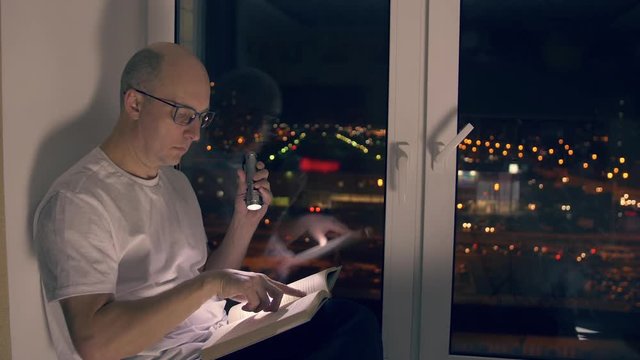 Adult man sitting on windowsill and reading book with flashlight in night