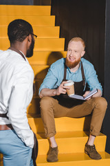 two multiethnic male baristas looking at each other and discussing work on stairs