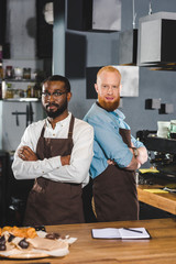 two young multicultural male baristas in aprons standing with crossed arms in coffee shop