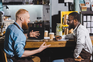 smiling young multicultural owners of coffee shop talking and sitting at table with laptop and coffee cups