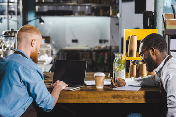 young male multiethnic owners of coffee shop working at table with textbook and laptop