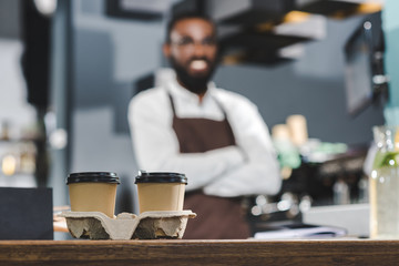 Fototapeta na wymiar close-up view of two paper cups with coffee and smiling african american barista standing with crossed arms behind