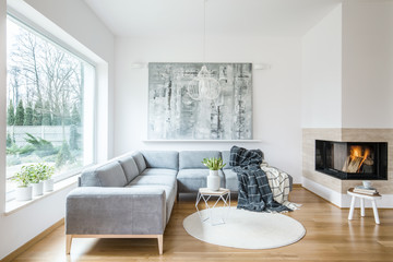 White sitting room interior with corner grey sofa, tulips in vase placed on end table, fireplace and modern art painting - obrazy, fototapety, plakaty