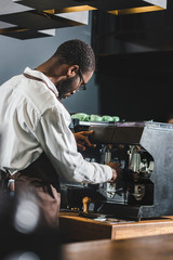 young african american barista in eyeglasses and apron working with coffee machine