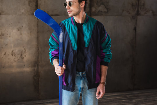 stylish young man in vintage windcheater and sunglasses with hockey stick looking away