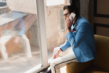 Fototapeta premium handsome young man in stylish suit talking by phone while sitting on couch