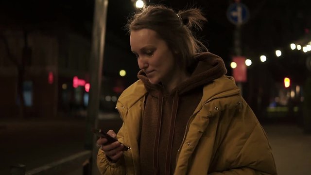 Short haired girl walks through the evening city smiling, stops and texting in the smartphone. Handheld footage