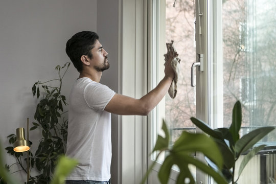 Side view of young man cleaning glass window in living room