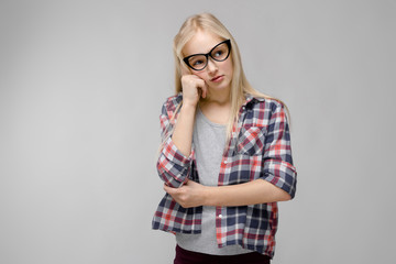 Portrait of attractive sad sweet adorable blonde teenager girl in checkered clothes in glasses holding hand near her face on gray background