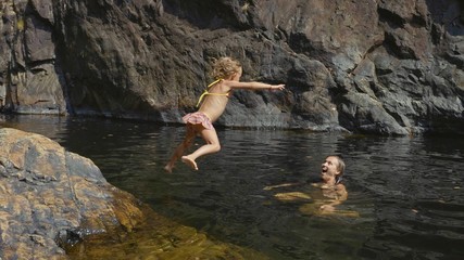 Little cute girl jumping in water of mountain lake to her mother