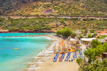 Sunny sandy Livadi beach in sea Bay of resort village Bali. Views of mountain, shore, washed by waves and sun loungers with parasols where sunbathing tourists. Bali, Rethymno, Crete, Greece