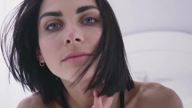 Portrait of young sexy Caucasian brunette in black lingerie, looking at camera, gently touching her neck, blinking 50 fps