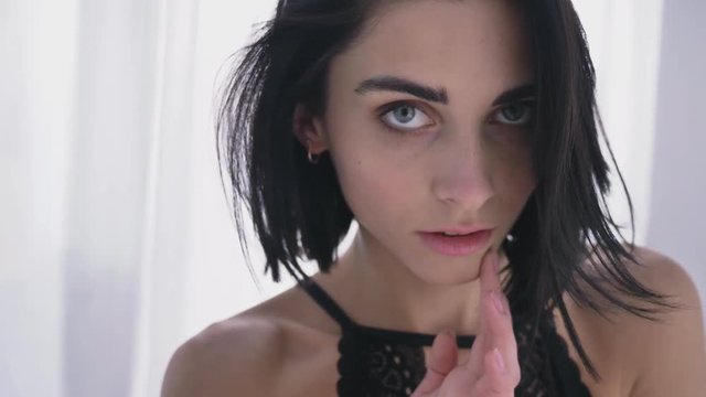 Portrait of young sexy caucasian brunette in black lingerie, looks down, gently touches her face, blinks 50 fps