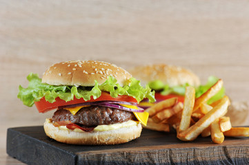 Cheeseburger from chopped beef cutlet with tomato, salad, bacon, sauce and red onion. Next to the cheeseburger is French fries. In the background is another cheeseburger. Light background. Close-up. 