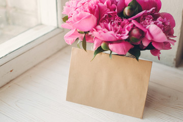 lovely pink peonies bouquet with empty craft card on rustic white wood at old window in light, top view, space for text. floral greeting card mock-up. happy mothers day concept