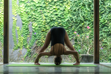 fit young woman practicing yoga in Forward Bend pose (Uttanasana) in front of wall covered with green leaves