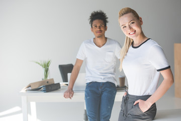 beautiful young woman smiling at camera while standing with african american colleague in office