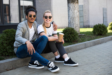 happy young multiethnic couple smiling at camera while sitting on street with coffee to go