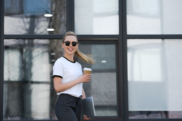 beautiful young woman in sunglasses smiling at camera while holding paper cup and laptop outside office building