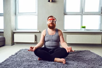 Fotobehang A funny fat bearded man in the headphones does yoga in the room. © Studio Romantic
